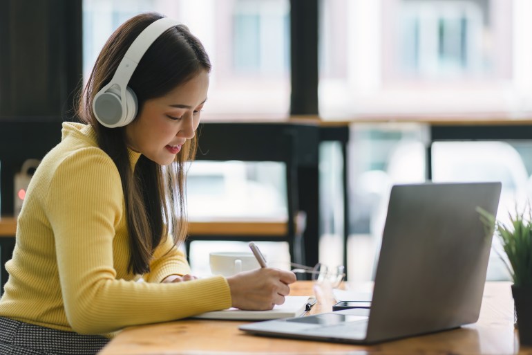 Focused Asian businesswoman wearing headphones is taking notes in a notebook while watching a webinar video course. Serious female student listening to the lecture to study online through e-learning