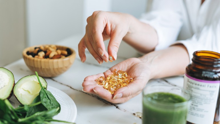 Woman doctor nutritionist hands in white shirt with omega 3, vitamin D capsules with green vegan food. The ...