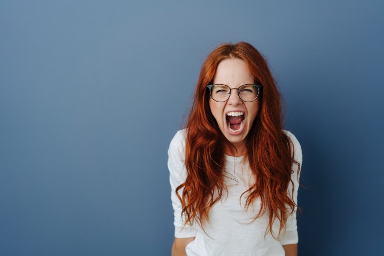 Angry young woman throwing a temper tantrum yelling at the camera with a furious expression over a blue studio background with copy space