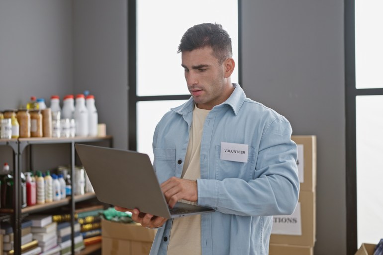 Devoted young hispanic man volunteering at community charity center, dutifully working with laptop amidst donation boxes,; Shutterstock ID 2426565675; purchase_order: aljazeera ; job: ; client: ; other: