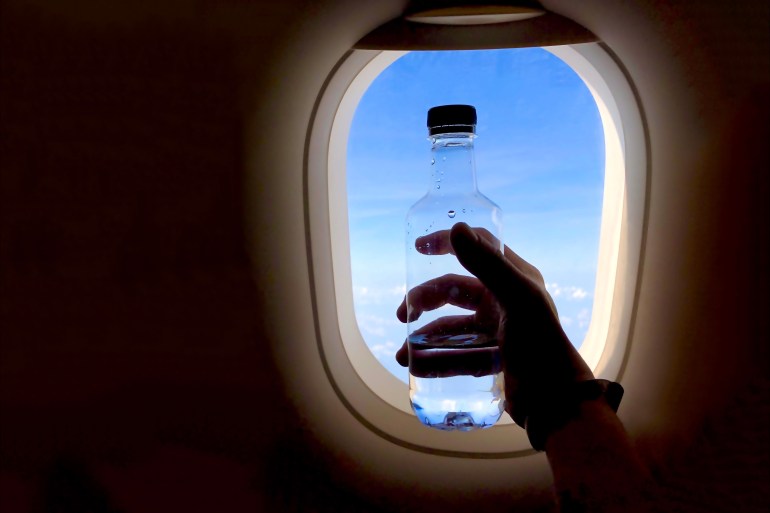 hand holding water bottle near airplane window; Shutterstock ID 1627184908; purchase_order: aljazeera ; job: ; client: ; other: