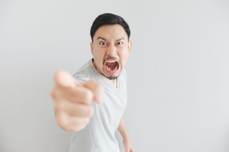 Angry face of Asian man in grey t-shirt with hand point on empty space. المصدر: ادوبي ستوك